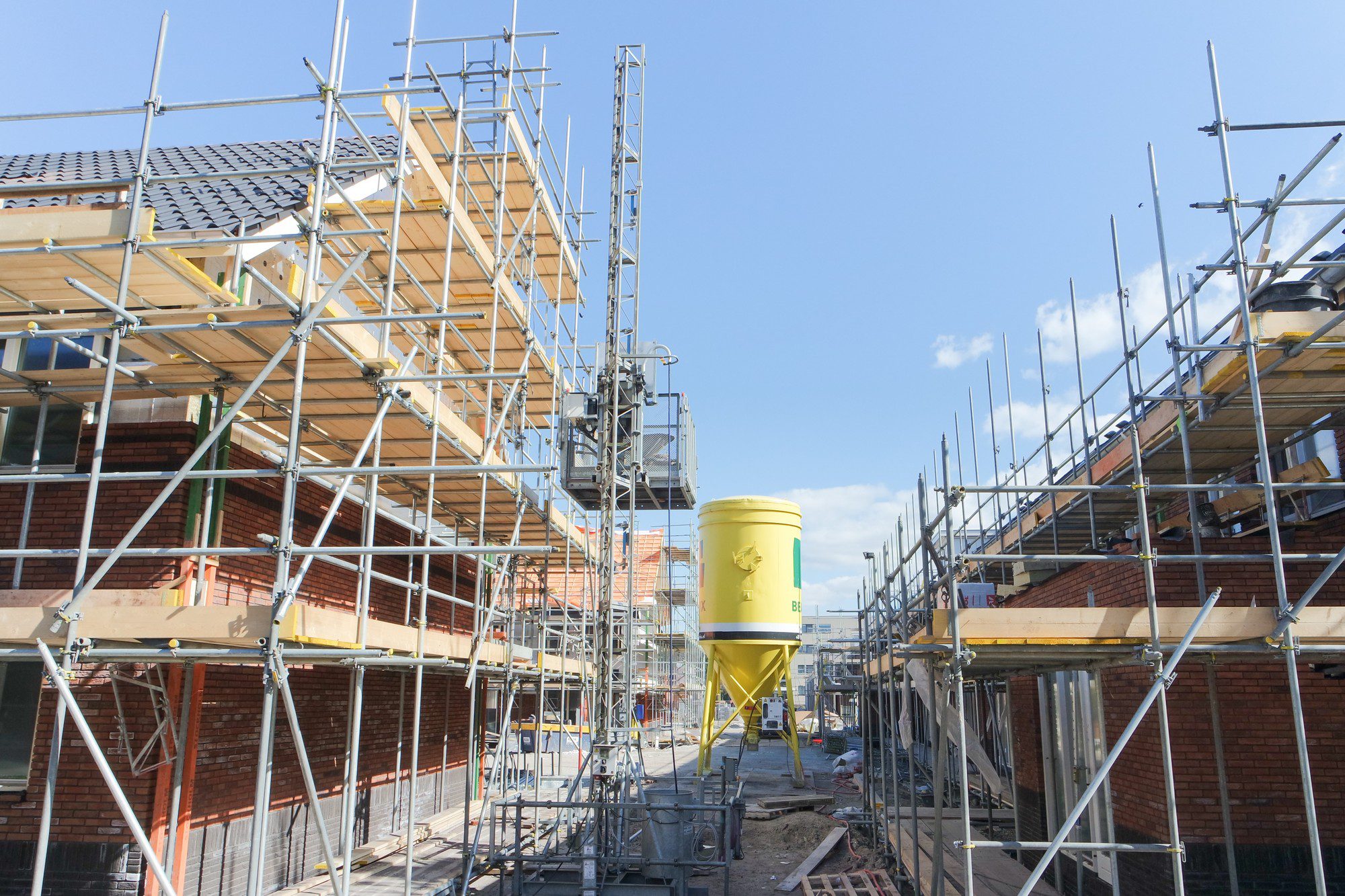 The image shows a construction site with houses in the process of being built. There is an extensive scaffolding structure in place around the partially constructed buildings, which consist of brick walls and wooden roof structures. The roof tiles have been installed on some parts of the roofs. In the foreground, there's a large yellow construction container, likely used for storing construction materials or waste. The sky is partly cloudy, suggesting fair weather conditions for construction work. The area appears to be neatly organized, indicating a well-managed construction site.