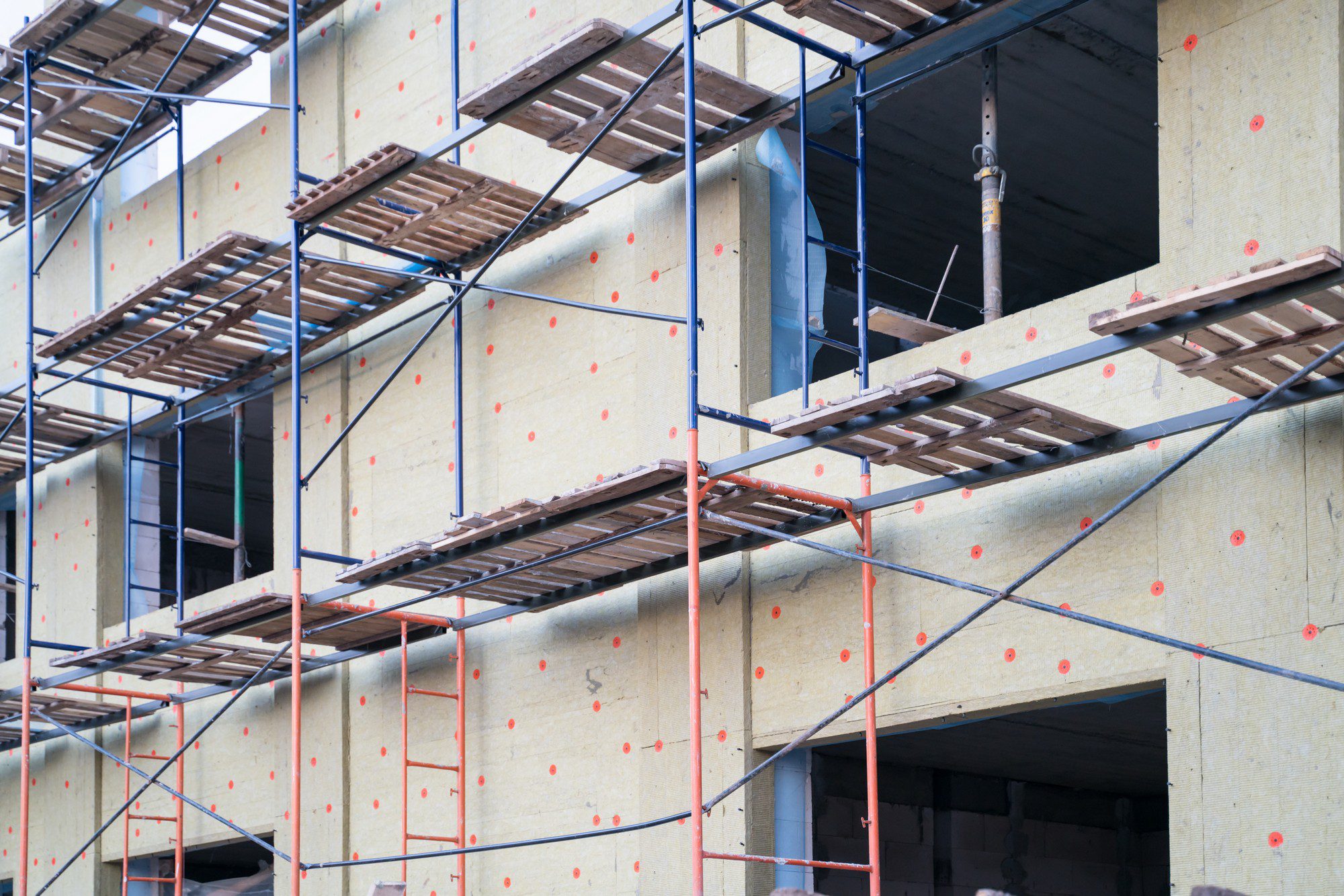 The image shows a section of a building under construction, featuring scaffolding structures set up along its exterior wall. Scaffolding is composed of metal pipes and connectors, with wooden planks laid across for workers to stand on. The wall itself appears to be insulated, as indicated by the yellow material with red spot markers which could be for fastening the outer layer or for reference points in the construction process. There are visible windows without glass installed yet. Overall, the image captures the ongoing work and structure of a building in the middle of being built or renovated.