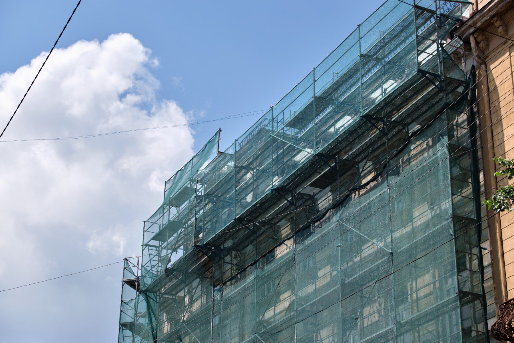 The image shows a building covered with scaffolding and green safety netting, which indicates that construction, renovation, or repair work is taking place. The scaffolding comprises several levels of metal structures to support workers and materials. The green netting is typically used to catch debris and ensure the safety of people below. Overhead there are some power lines, and the sky is mostly clear with a few clouds. A small portion of a tree can be seen on the lower right corner of the image, adding a touch of greenery to the urban environment.