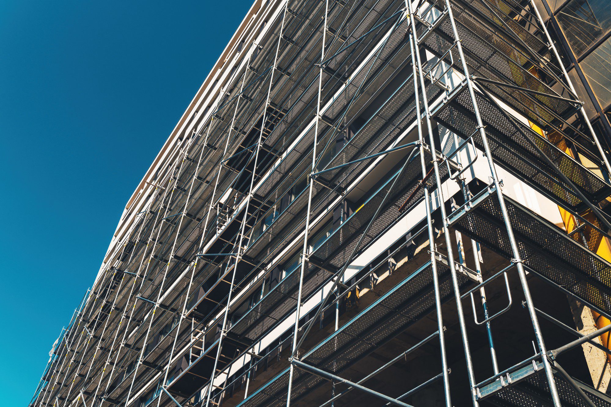 The image shows a structure covered with scaffolding. The scaffolding consists of a network of metal pipes and planks, which are used to provide a temporary platform for workers and materials during the construction, maintenance, or repair of buildings and other structures. The scaffolding covers the facade of the building, and it appears to be a multi-story structure given the number of levels of scaffolding present. The sky is clear and blue, indicating good weather conditions. The sunlight casts shadows on the building, highlighting the geometric pattern of the scaffolding.