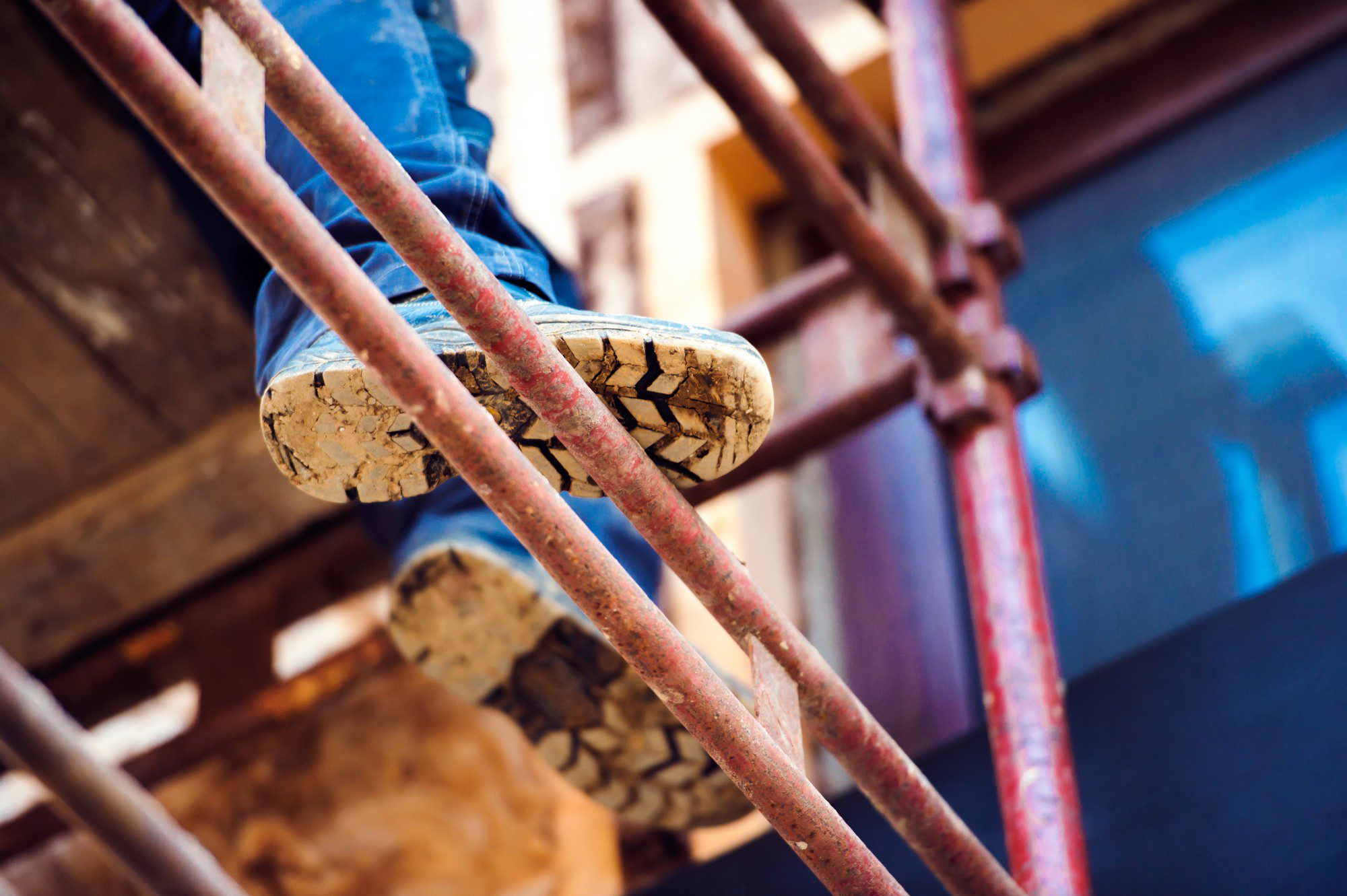 In the image, there's a close-up shot of a person's feet as they climb up a metal scaffold. The person is wearing rugged work boots that have a noticeable amount of dirt on the soles, indicating they have been used on a construction site or in a similar outdoor setting. The scaffolding itself shows signs of wear and rust, hinting at its outdoor use. The focus on the boots and scaffold suggests the image's emphasis on construction work, safety, and the physical aspect of labour-intensive jobs. The background is out of focus, but there seems to be a building structure behind.