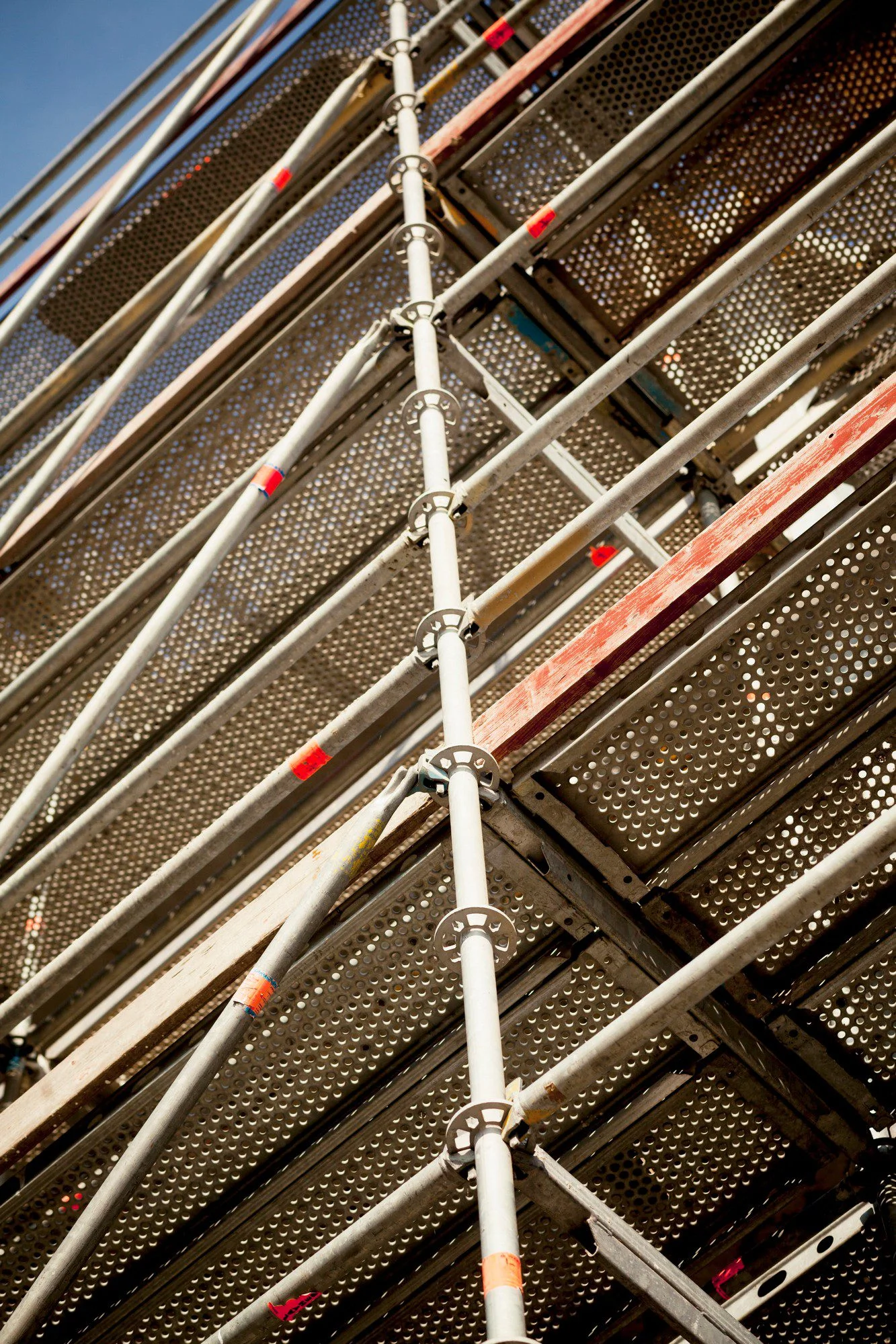 The image shows a close-up view of a section of scaffolding. It features several vertical and horizontal metal poles connected with circular clamps and diagonal bracing for stability. The metal platform decks, which provide a working surface, have a perforated pattern for grip and are visible in the background. Red and white markings, possibly for safety or identification, can be seen on some of the poles. The scaffolding is typically used in construction or maintenance work to provide temporary access to higher levels of a building or structure.