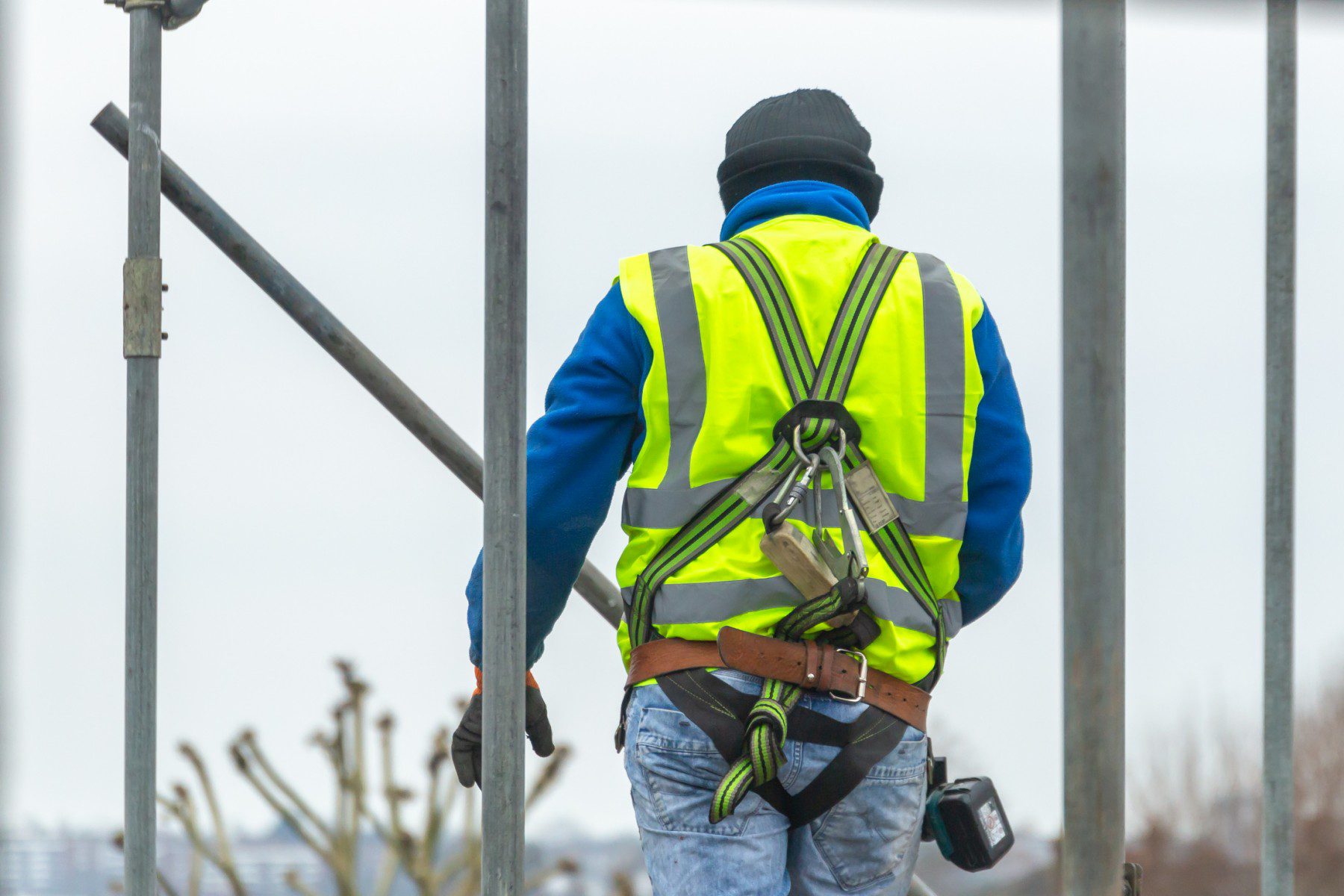 This image depicts a construction worker from behind. The worker is wearing a high-visibility safety vest with reflective stripes, which is typically used to ensure that personnel are visible on construction sites or in other potentially hazardous working environments. He also has on a blue long-sleeve shirt, a black beanie, gloves, and jeans. The worker is equipped with a safety harness that has a lanyard attached to it to prevent falls, indicating that he might be working at a height. There is a tool belt around his waist, and a hand-held radio is attached to his belt, which is possibly used for communication on the site. The construction scaffolding around the worker indicates that the photo was taken on an active construction site.