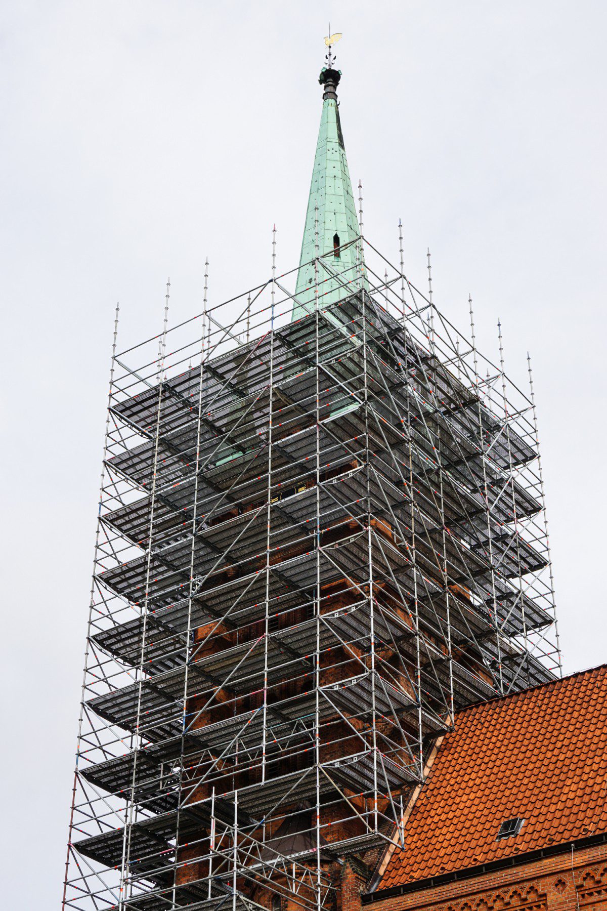 The image shows a church steeple that is under renovation or restoration. The steeple is covered with scaffolding, which is a temporary structure used to support work crews and materials during the construction, maintenance, or repair of buildings. The scaffolding reaches up to the top of the steeple, where a gold-coloured weathervane is visible. The body of the church visible at the bottom has a red tiled roof, and the masonry appears to be made of brick. The overcast sky suggests a subdued lighting condition, which is common during cloudy weather.