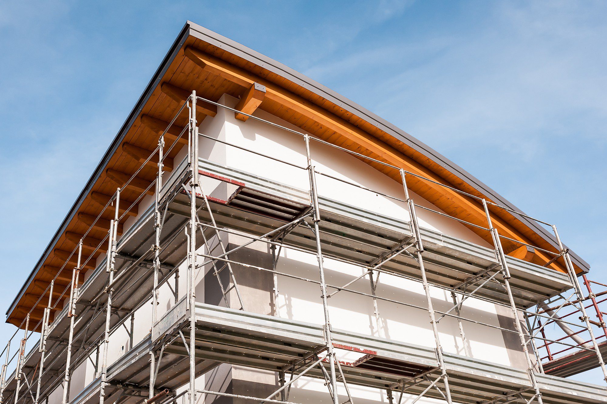 The image shows a building under construction or renovation, with scaffolding set up around it. The scaffolding consists of metal poles, cross braces, platforms, and ladders to allow workers access to the exterior walls of the building. The scaffolding surrounds a white building with a gray foundation and a wooden eave section, suggesting that work might be taking place on the exterior surfaces such as plastering, painting, or installing exterior finishes. The sky is visible in the background, indicating the photo was taken on a day with good weather. There are no visible people in the image.