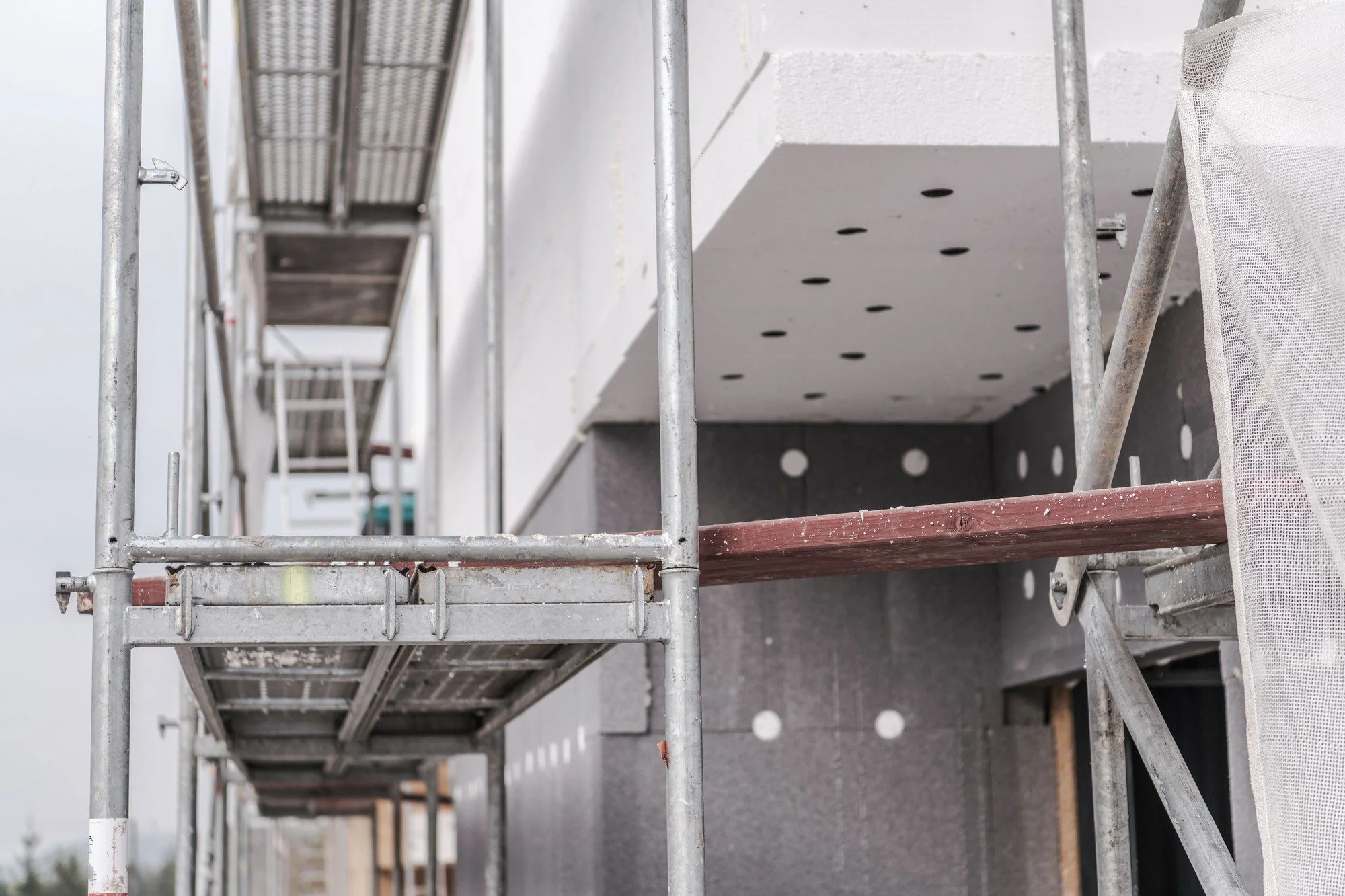 This image shows a section of scaffolding alongside a building under construction or renovation. The scaffolding is made of metal poles and has wooden boards laid across for workers to stand on. The side of the building features insulation panels that are typically used for thermal insulation, and there are holes for what might be eventual fixtures or ventilation. Over the insulation, there is a mesh covering, which is commonly used to hold insulation in place and provide a base for rendering or plastering. The environment looks overcast, which indicates the photo might have been taken on a cloudy day.